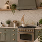 A styled kitchen scene with sage green cabinets, a matching range hood, a gas stove with brass knobs, a beige mosaic tile backsplash, and a decorative rabbit-shaped tile in the center. A white Dutch oven sits on the stove, with plants, jars, and wooden shelves around it.
