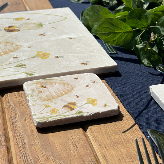 A wooden dining table set with cream marble placemats and coasters. The placemats are square and feature bees and a beehive in a buttercup meadow, in a watercolour style.