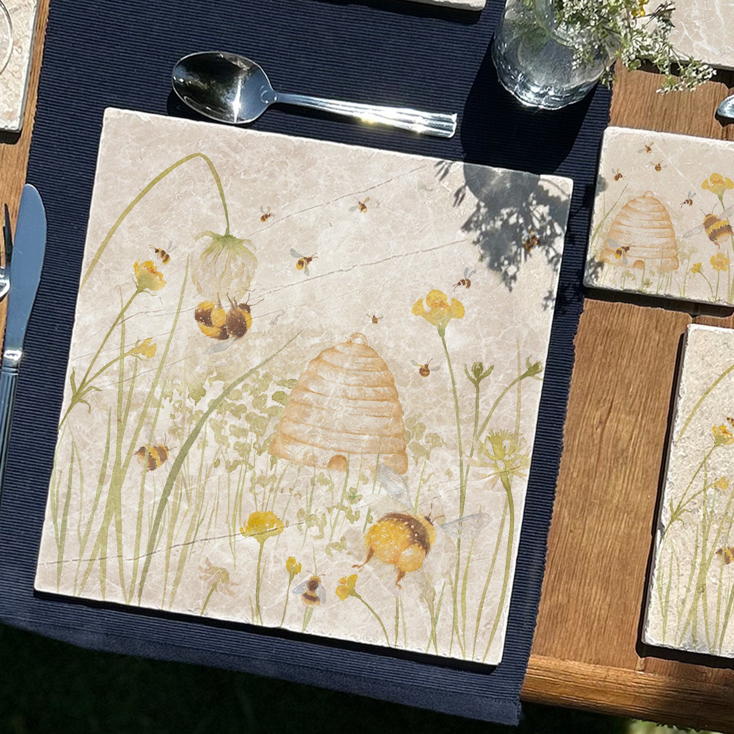 A wooden farmhouse dining table set with large square marble placemats. The placemats feature bees and a beehive in a buttercup meadow, in a watercolour style.
