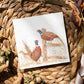 A greetings card laid flat on a table surrounded by gift wrapping items including scissors and ribbon. The card features a group of pheasants in a watercolour style.