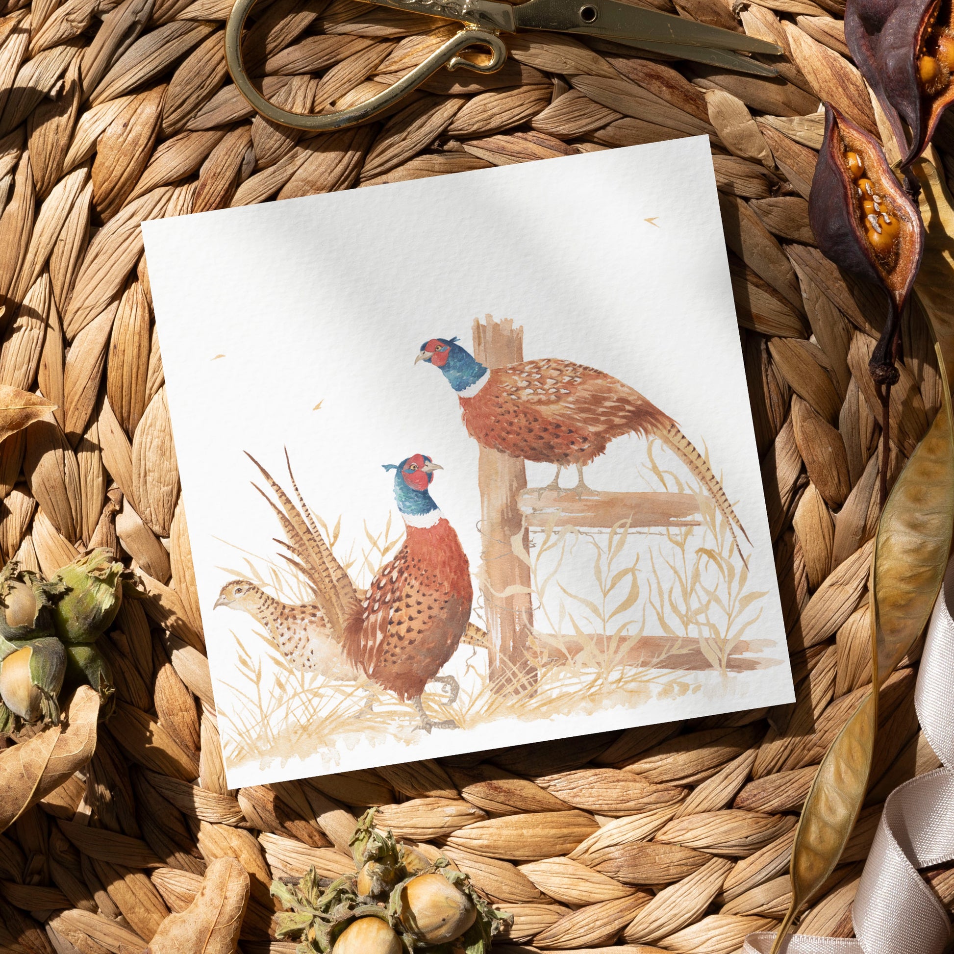 A greetings card laid flat on a table surrounded by gift wrapping items including scissors and ribbon. The card features a group of pheasants in a watercolour style.