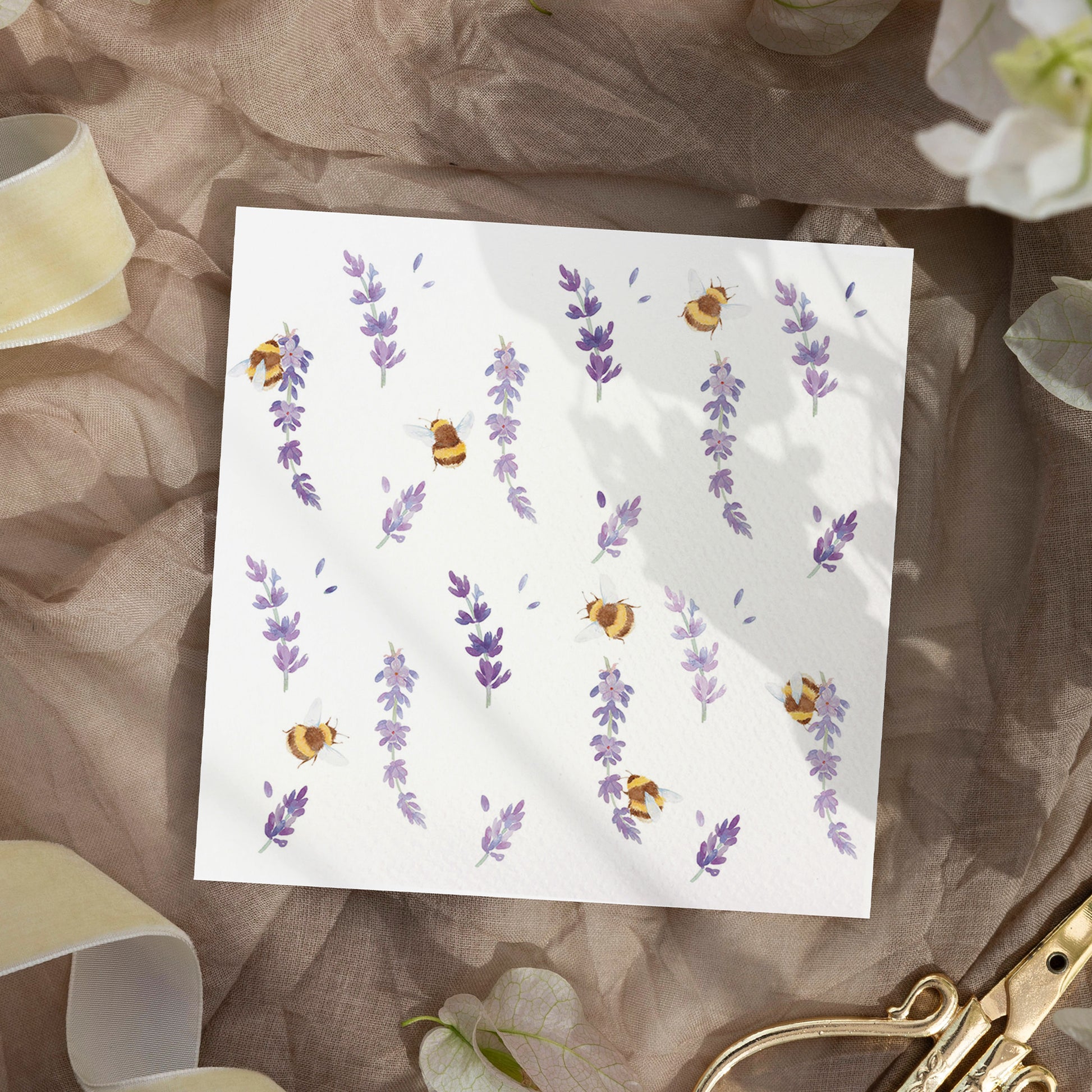 A square greetings card with a watercolour busy bee and purple lavender flowers pattern, laid on neutral fabric with ribbon and scissors.