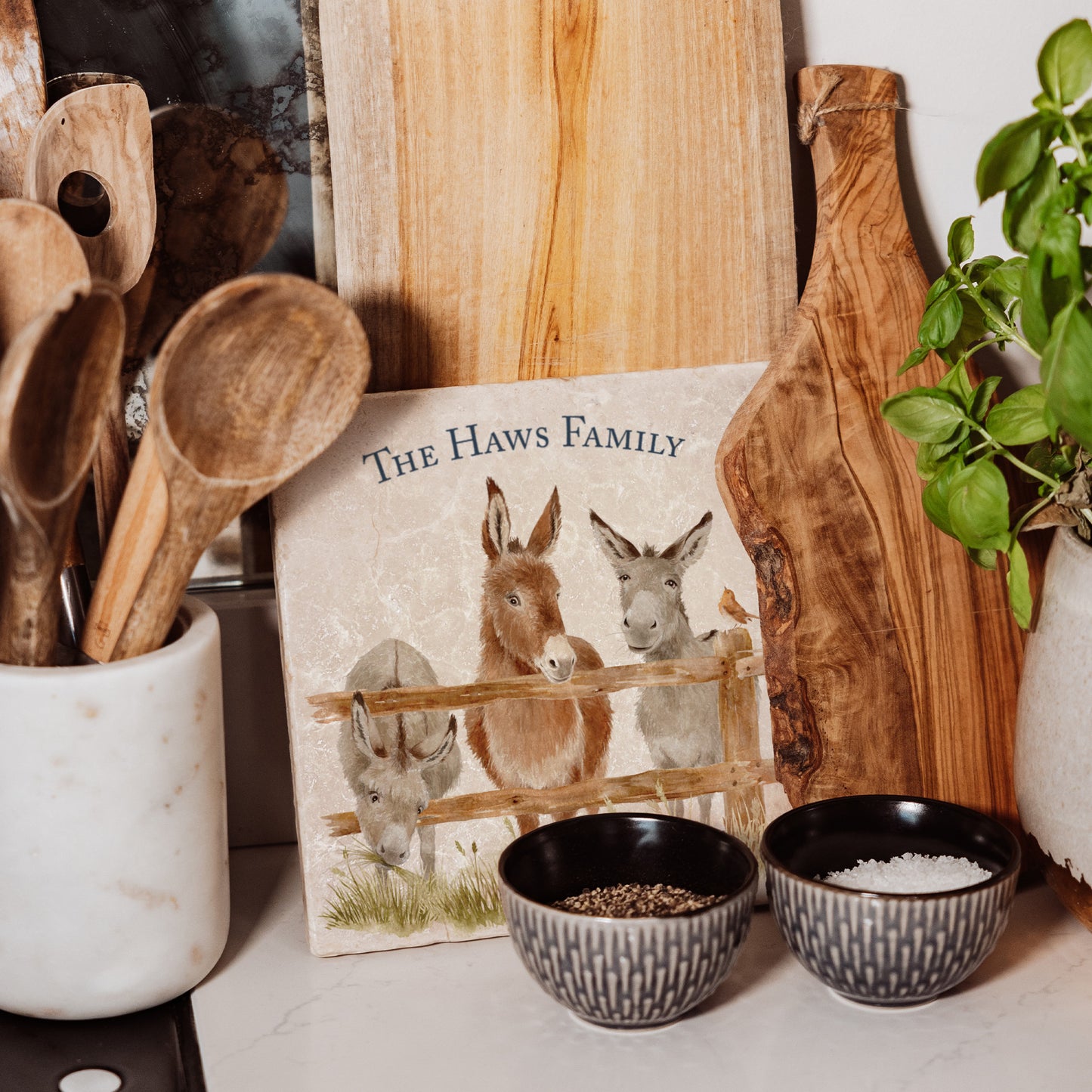 A handmade personalised medium marble platter featuring a watercolour design of three donkeys at a fence. The marble platter is leant up against a kitchen worktop with other wooden serving boards.