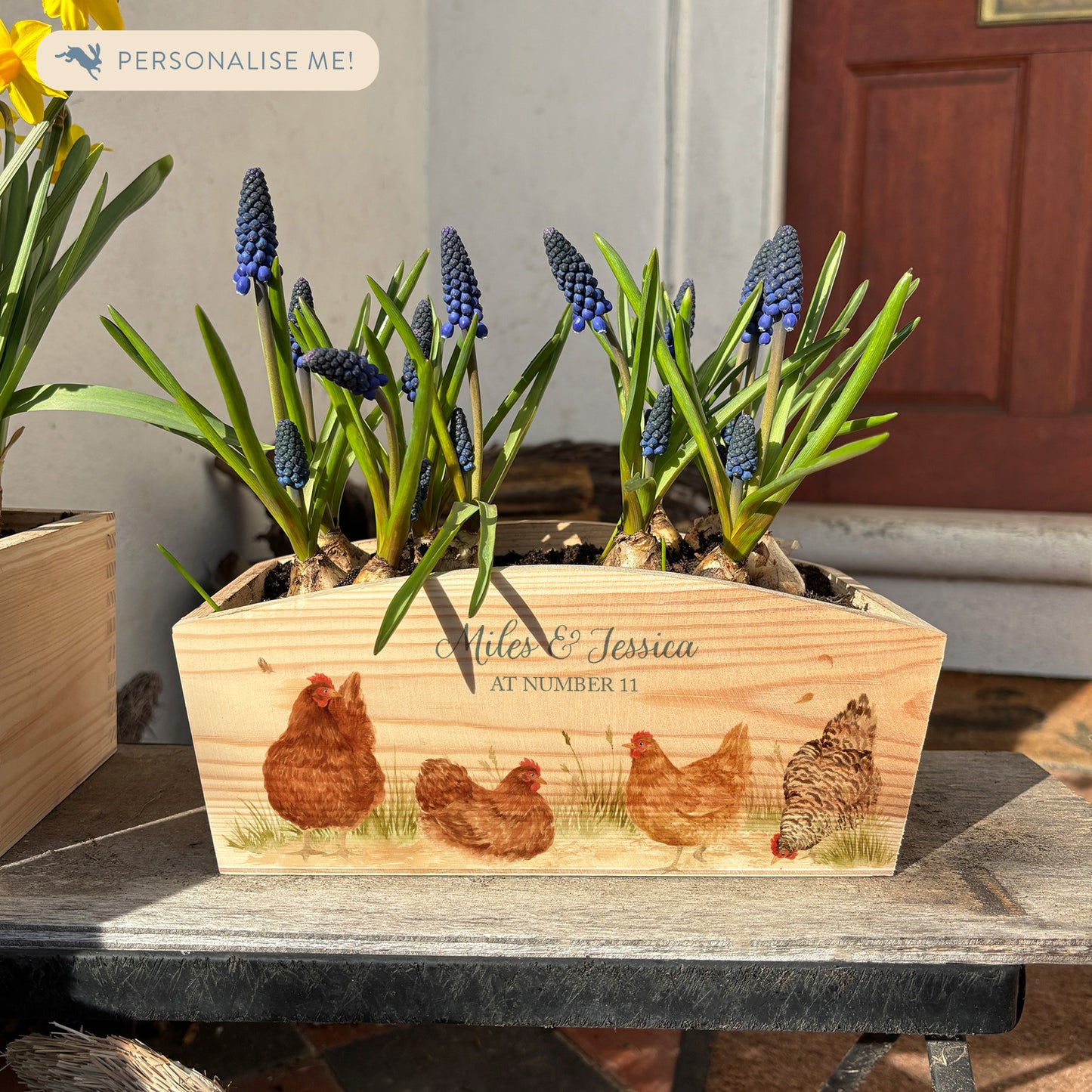 An arched wooden planter box full of spring bulbs. The planter box shows a watercolour design of a flock of hens pecking in the farmyard. The planter can be personalised with names and a selection of phrases, making it an ideal gift for any occasion.