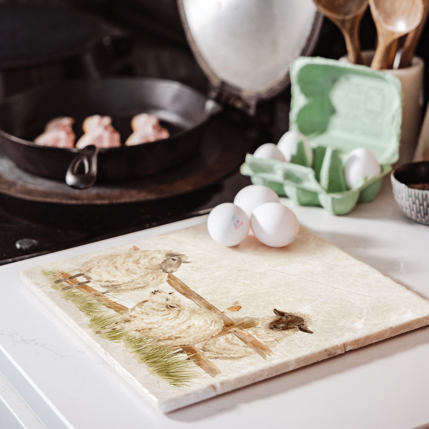 A large marble platter featuring a watercolour design of three sheep escaping from their field through a broken fence. The platter is set on a kitchen work surface next to the hob.