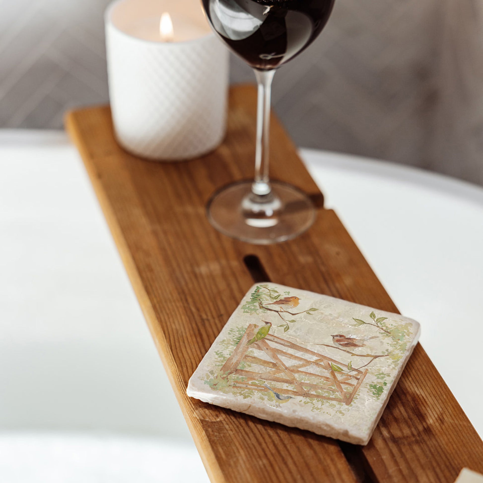 A square marble coaster on a wooden bath tidy above a bubble bath. The coaster features a watercolour design of garden birds in the hedgerow around a paddock gate.