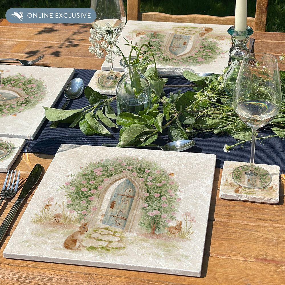 A handmade large marble placemat with a magical watercolour scene of a hidden garden door under an archway of roses. The placemat is set on a beautiful garden dining table with matching coasters.