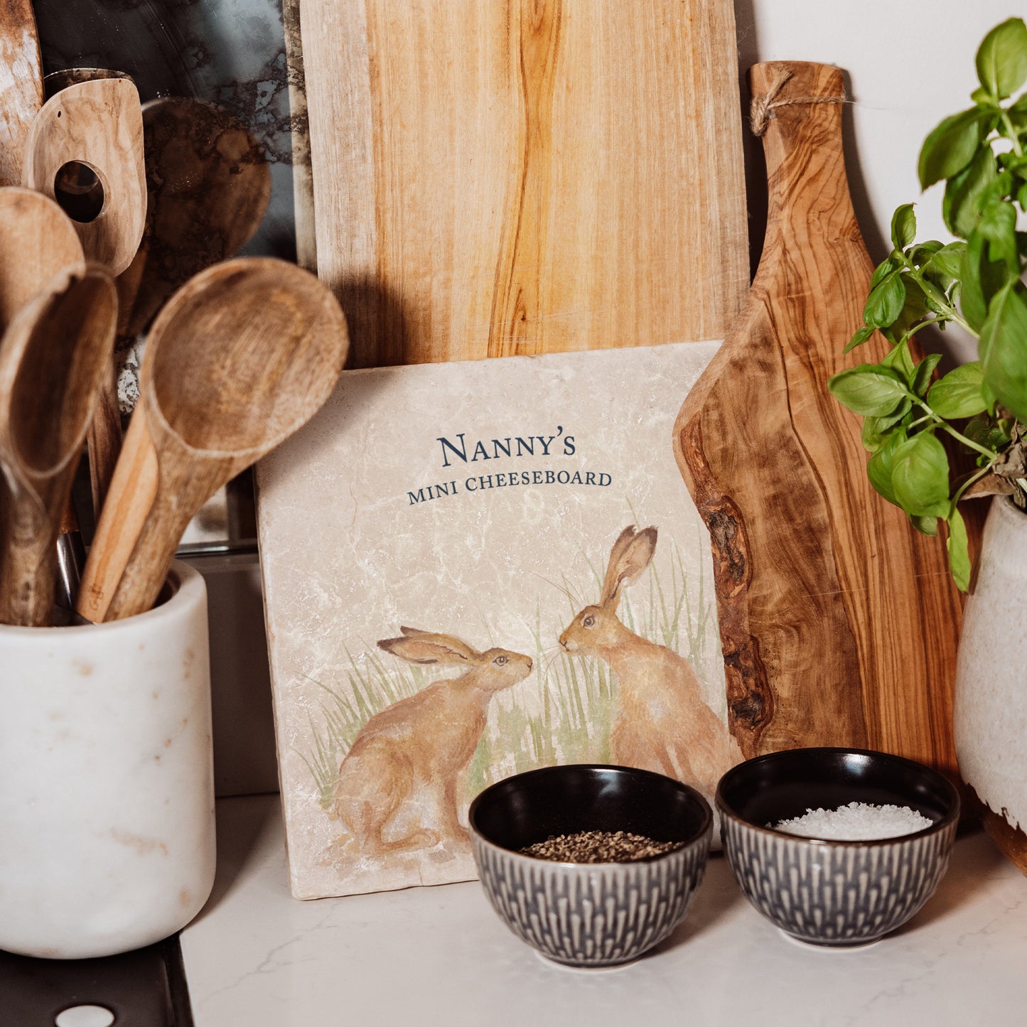 A medium personalised square cream marble platter propped up on a country kitchen worksurface among kitchen utensils and wooden chopping boards. The marble platter features a watercolour design of two hares facing each other about to touch noses.