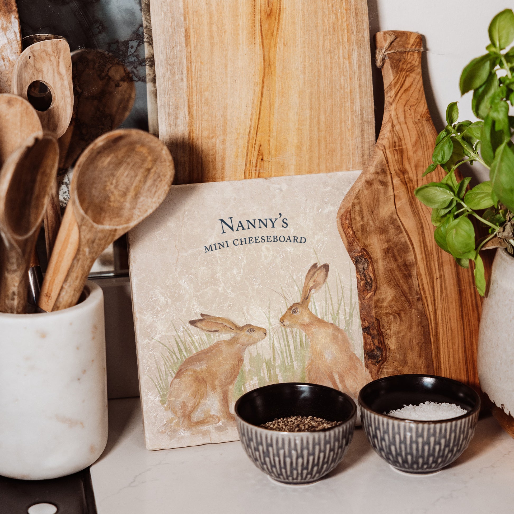 A medium personalised square cream marble platter propped up on a country kitchen worksurface among kitchen utensils and wooden chopping boards. The marble platter features a watercolour design of two hares facing each other about to touch noses.