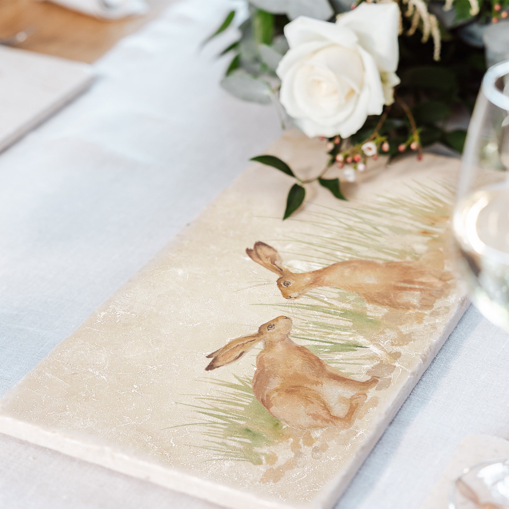 A marble sharing platter with a pair of hares watercolour design. The platter is set on a dining table to be used as trivet to serve hot pots and dishes onto.