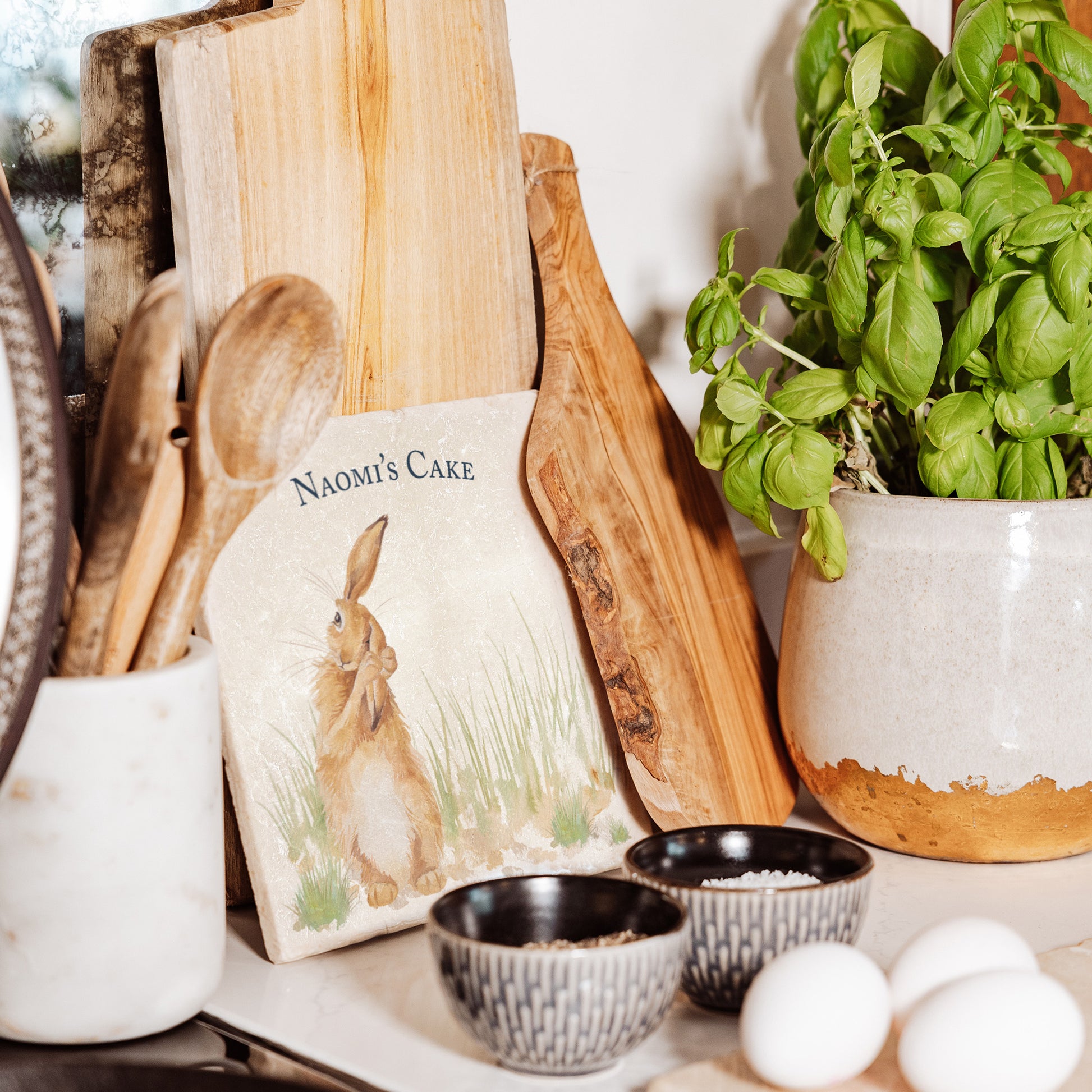 A medium square cream marble platter  which is personalised. It is propped up on a country kitchen worksurface among kitchen utensils and wooden chopping boards. The marble platter features a watercolour design of a hare washing his ear. 