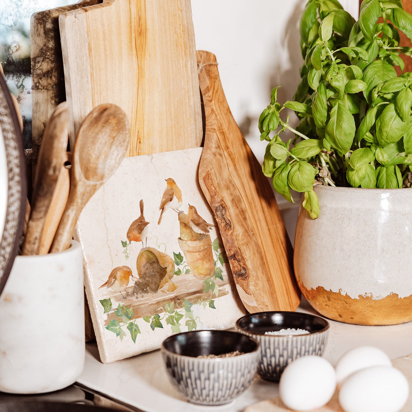 A medium square multipurpose marble platter, featuring a watercolour robin design. The platter is propped up on a kitchen worksurface ready to be used as a trivet.