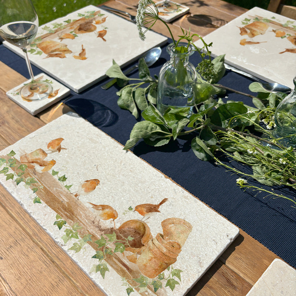 An outdoor garden table set with marble platters and coasters, each featuring a watercolour design of robins up to mischief on a shelf full of plant pots.