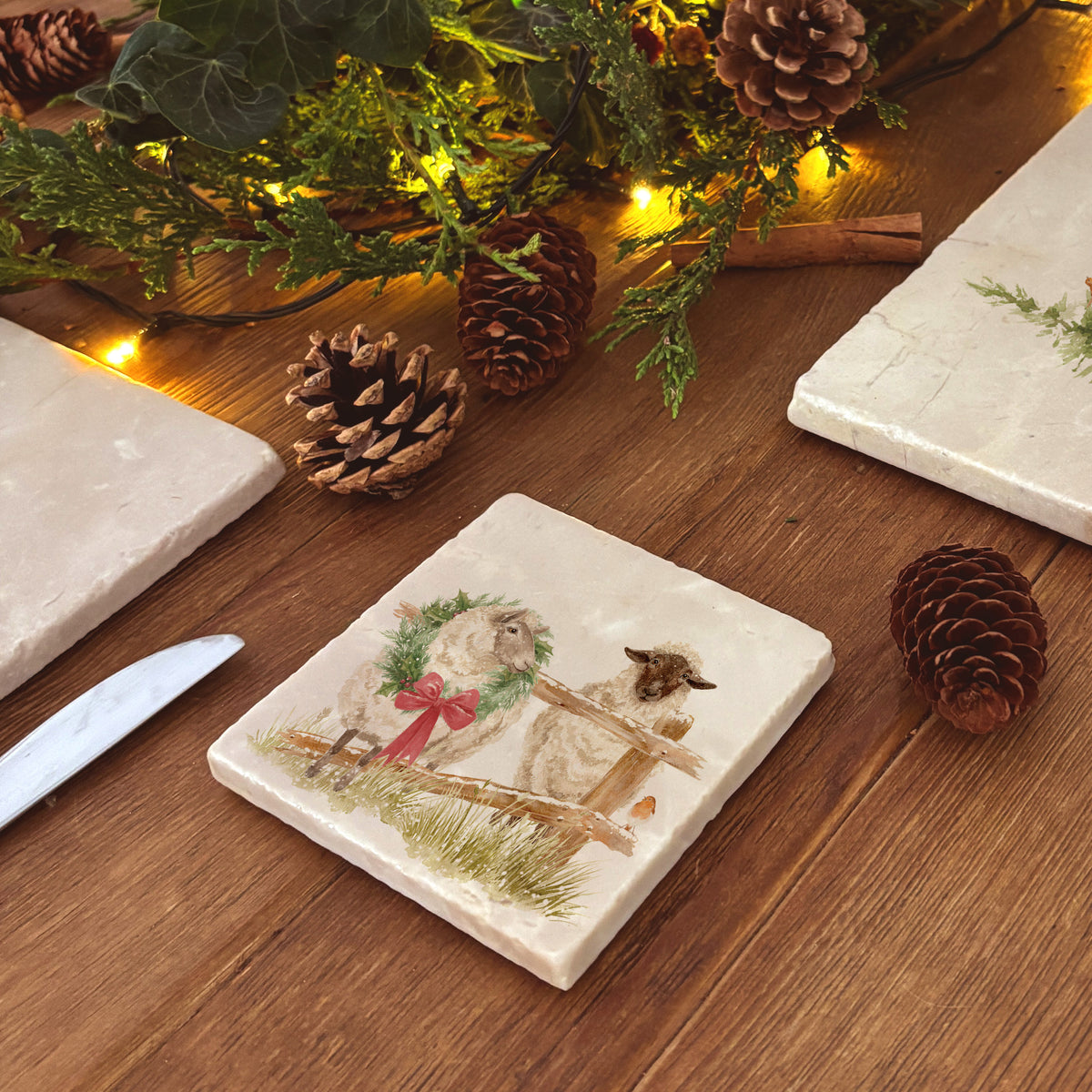 A set of four square marble coasters with a watercolour illustration of a farmyard scene, with sheep in the snow wearing a Christmas wreath.