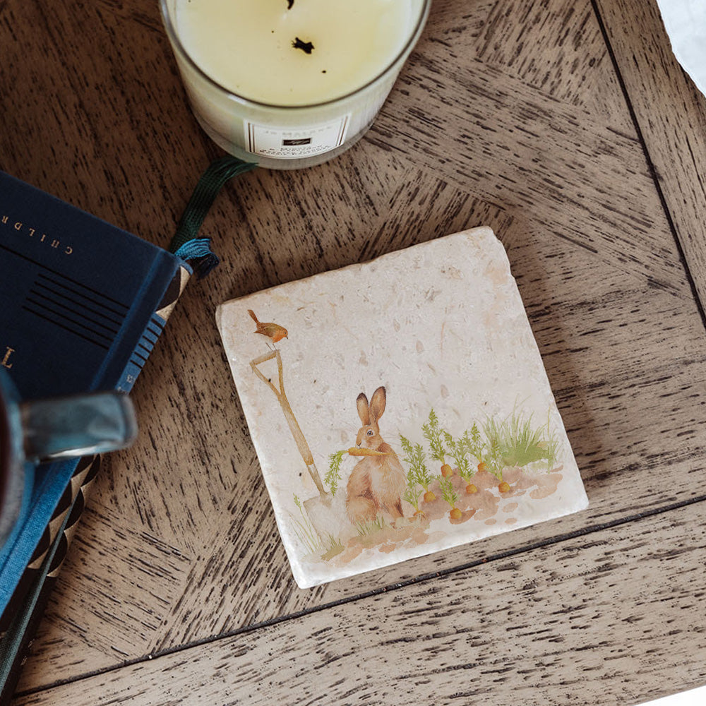 A marble coaster featuring a vegetable patch watercolour design with a hare eating carrots and a robin. The coaster is on a bedside table.