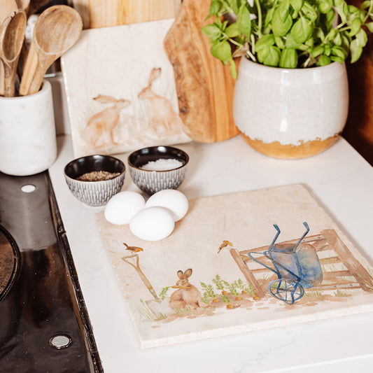 A large square handmade marble platter with a watercolour veg patch design. The marble platter is sat on a kitchen worktop next to the hob, ready to be used as a trivet.