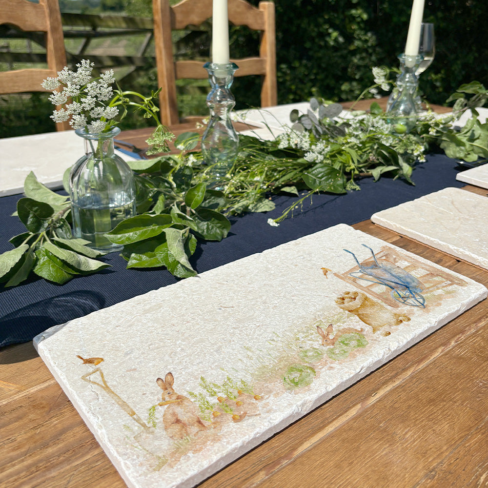 A garden table set with a marble sharing platter as a table runner. The platter has a watercolour vegetable patch design featuring hares eating the carrots and the robins keeping watch for the gardener.