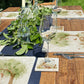 An outdoor dining table set with marble placemats and coasters featuring a watercolour scene of woodland animals under a willow tree.