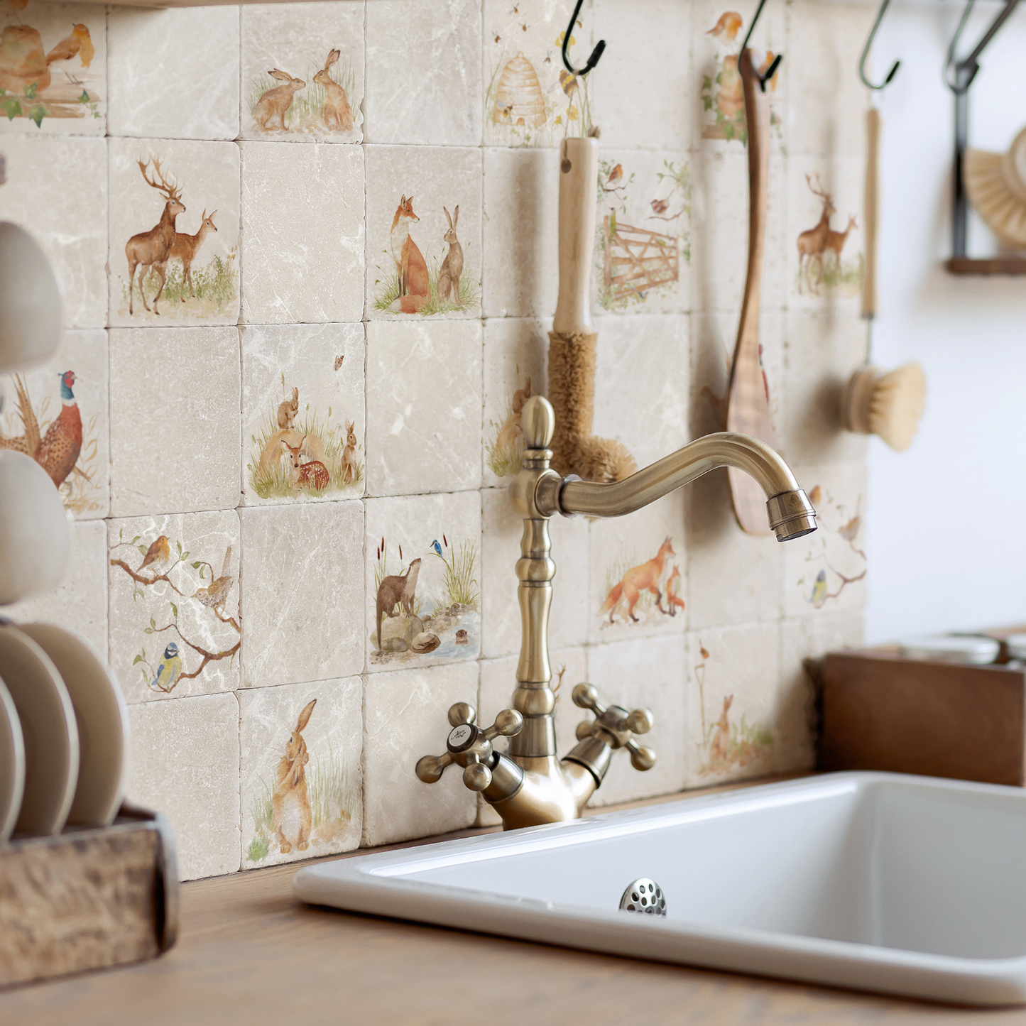 A rustic kitchen scene featuring a white sink with a vintage-style brass faucet mounted against a beige tiled backsplash decorated with watercolour animal illustrations. Wooden hanging utensils and a stack of white plates are visible in the background.