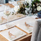 A wooden dining table set with cream marble placemats and coasters. The placemats are square and feature a hare washing his ear in a watercolour style.