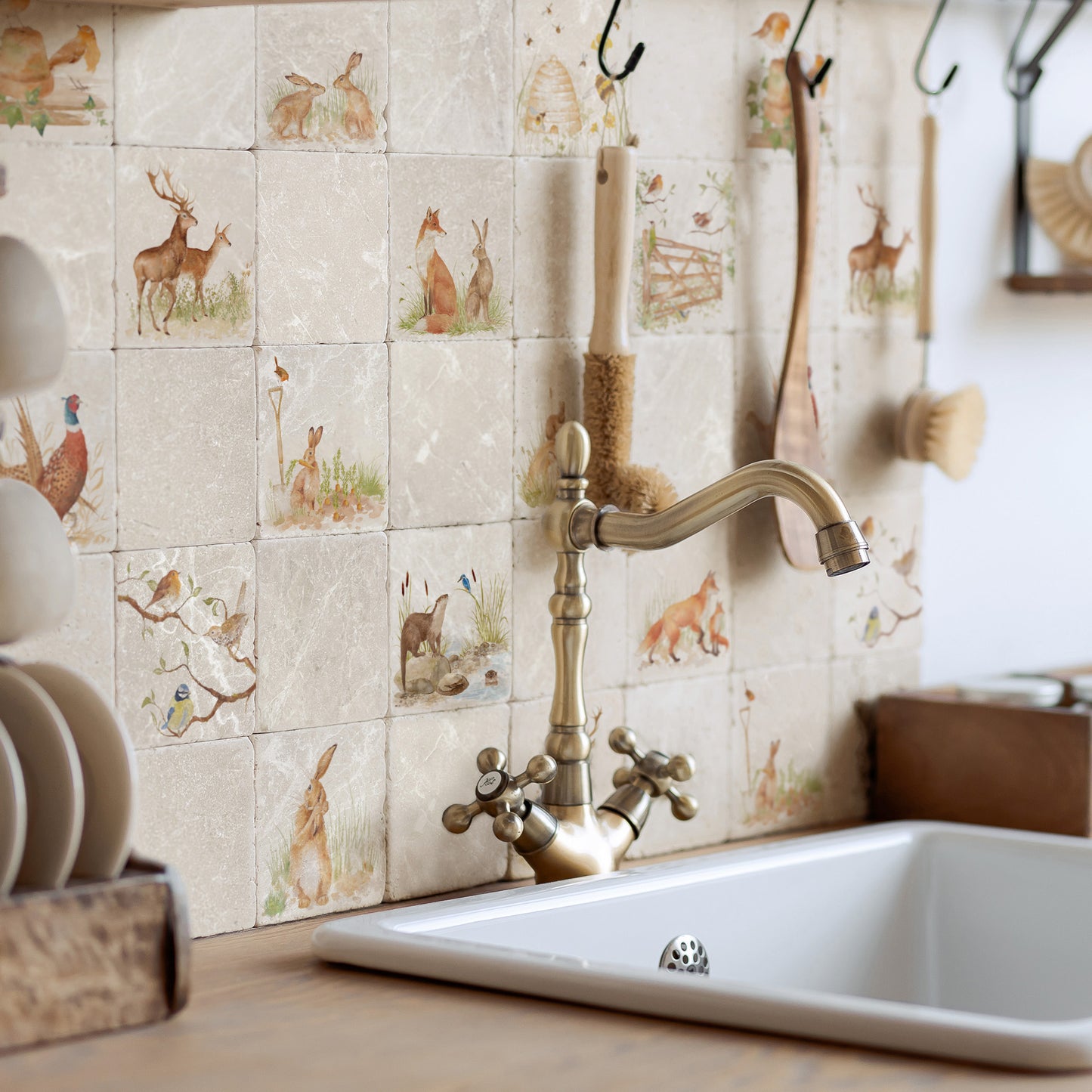 A kitchen backsplash made of square beige marble tiles with watercolour woodland animal illustrations, installed behind a brass faucet and a white sink. Wooden utensils hang from hooks on the wall, and a stack of white plates sits on the left.