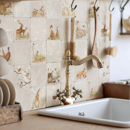 A kitchen backsplash made of square beige marble tiles with watercolour woodland animal illustrations, installed behind a brass faucet and a white sink. Wooden utensils hang from hooks on the wall, and a stack of white plates sits on the left.