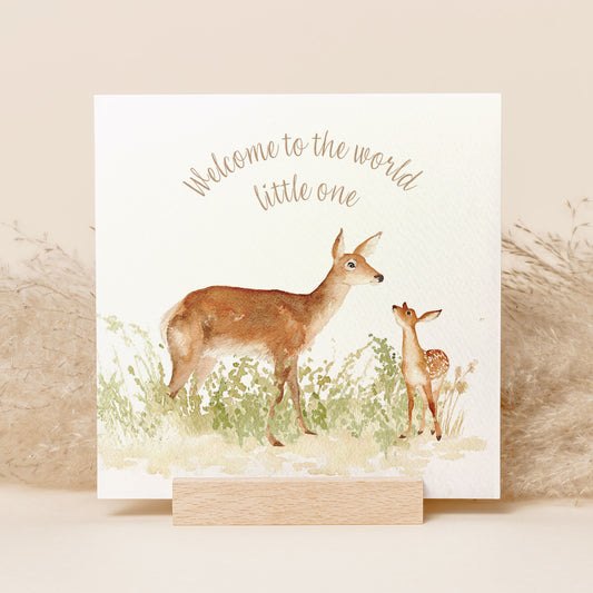A greetings card reading Welcome to the World Little One in brown text above a red deer doe and baby fawn in a watercolour style.