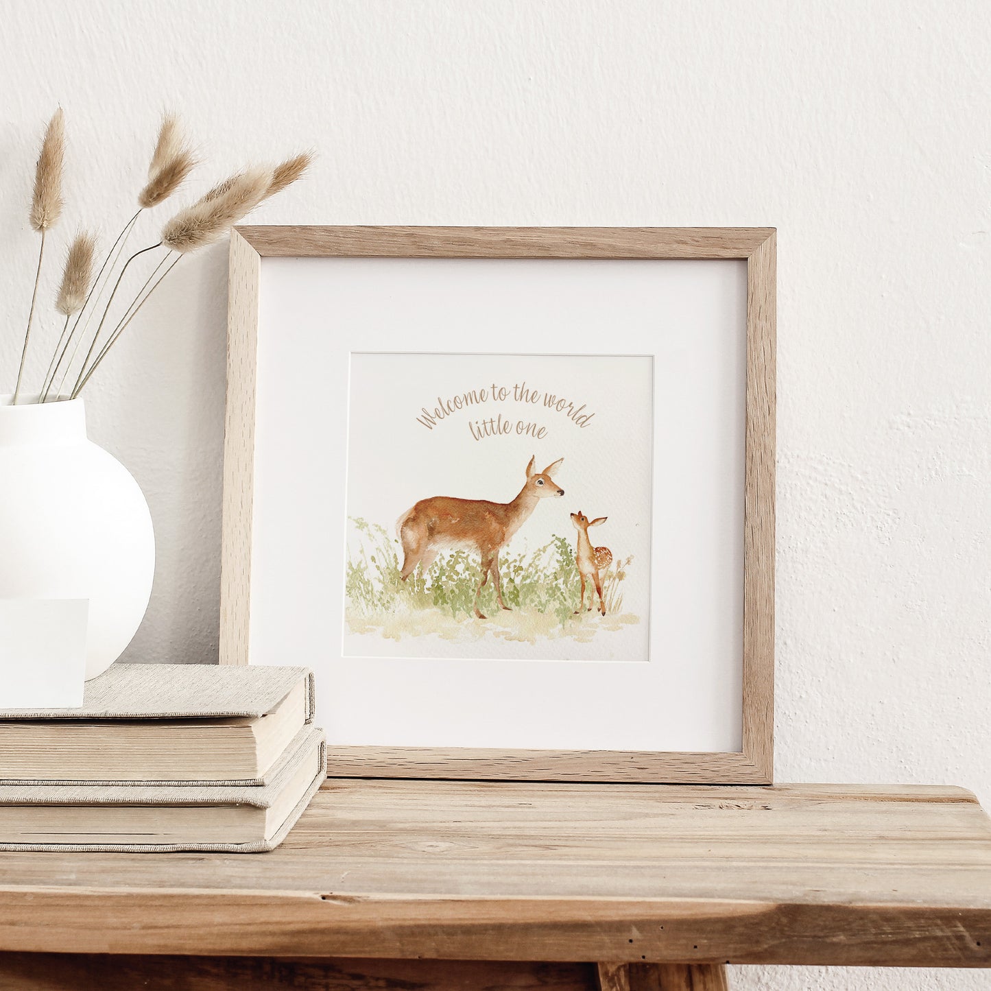 A greetings card displayed as an art print in a neutral coloured frame propped up on a bookshelf. The card reads Welcome to the World Little One in brown text above a red deer doe and baby fawn in a watercolour style. 