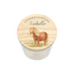 A personalised candle in a white cylindrical jar with a round wooden lid. The lid features an illustration of a brown pony with a blonde mane holding flowers in its mouth, with the text 'CONGRATULATIONS Isabella' written above it.