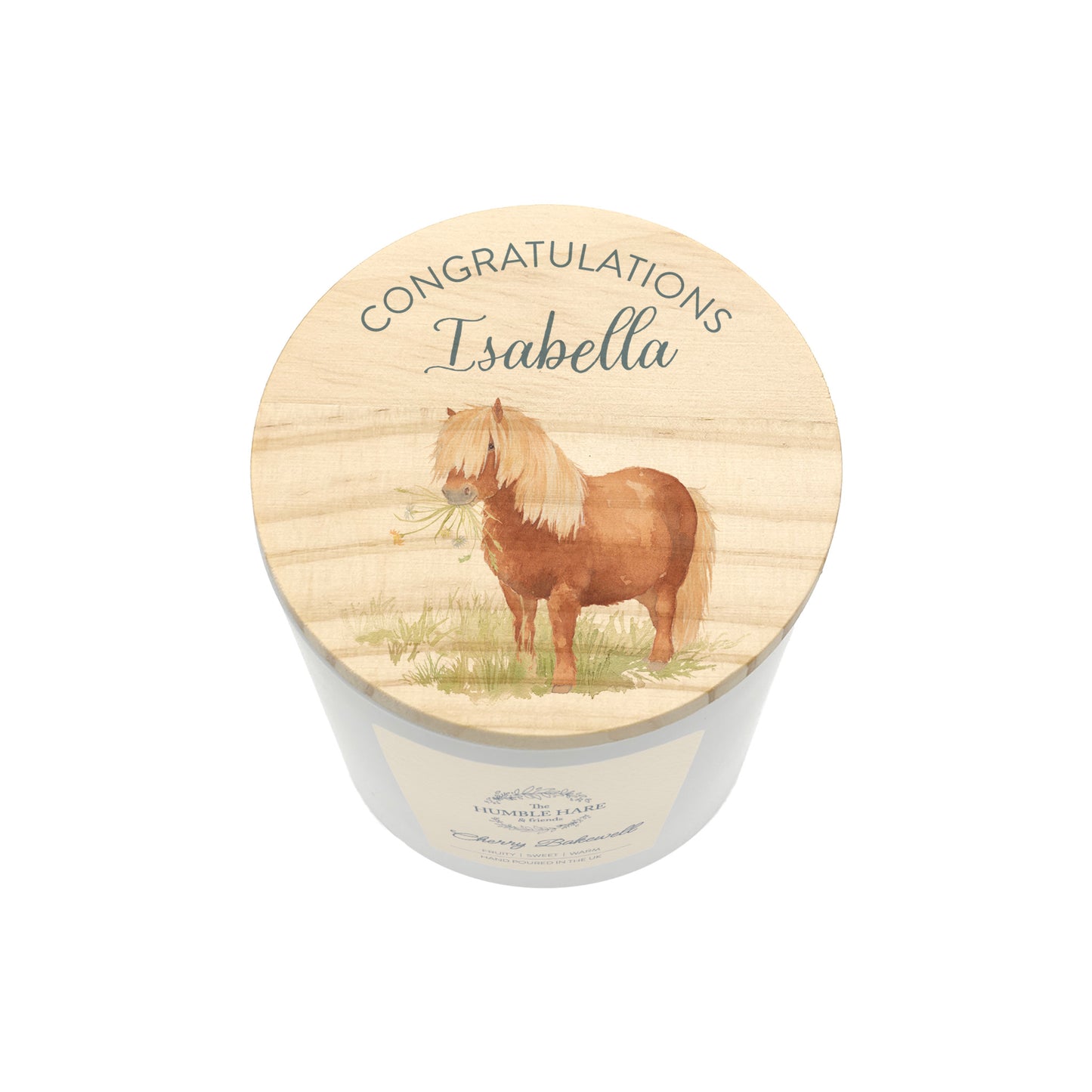 A personalised candle in a white cylindrical jar with a round wooden lid. The lid features an illustration of a brown pony with a blonde mane holding flowers in its mouth, with the text 'CONGRATULATIONS Isabella' written above it.