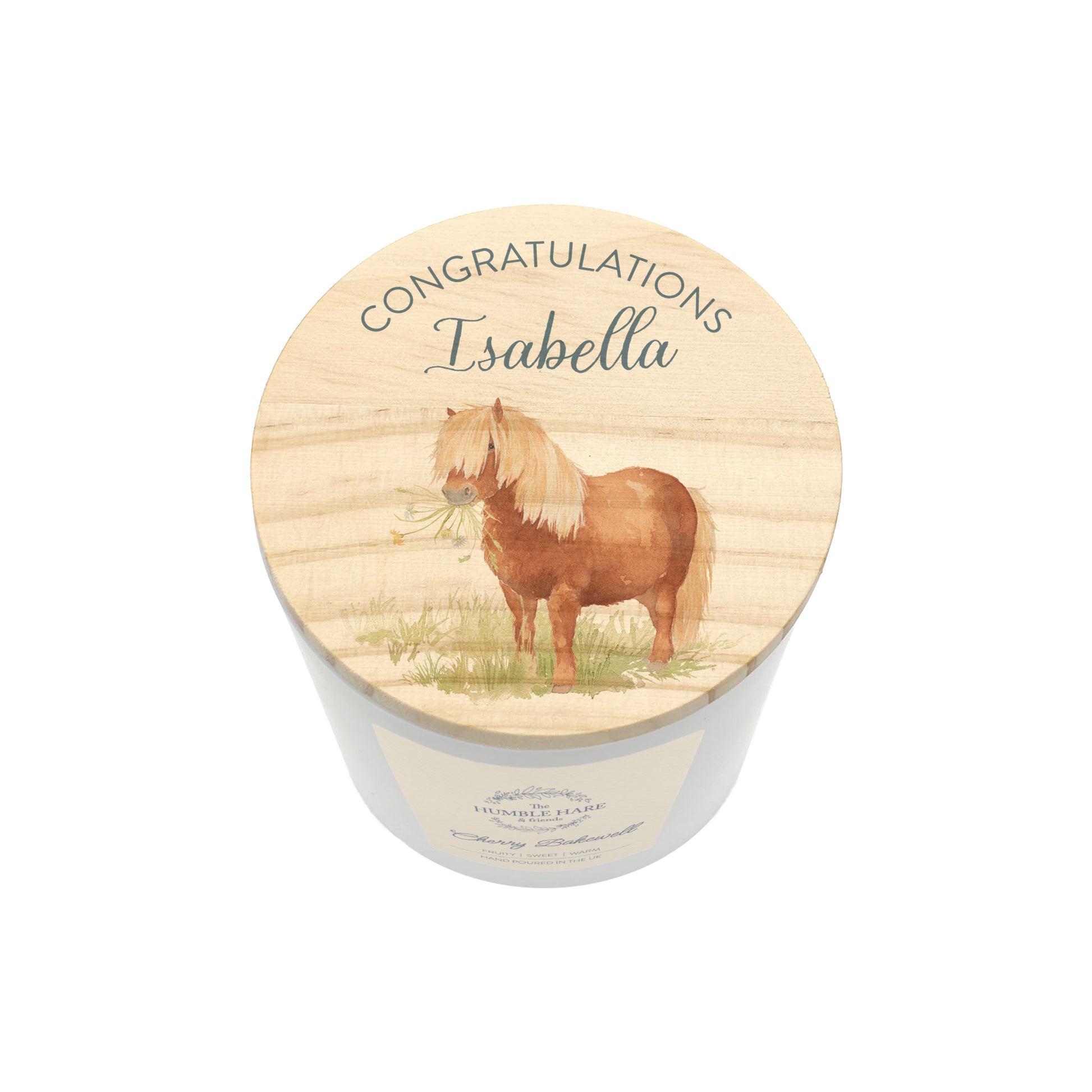 A personalised candle in a white cylindrical jar with a round wooden lid. The lid features an illustration of a brown pony with a blonde mane holding flowers in its mouth, with the text 'CONGRATULATIONS Isabella' written above it.