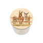 A cylindrical white candle jar with a round wooden lid with a watercolour illustration of three donkeys behind a fence and a robin, shown on a plain white background.