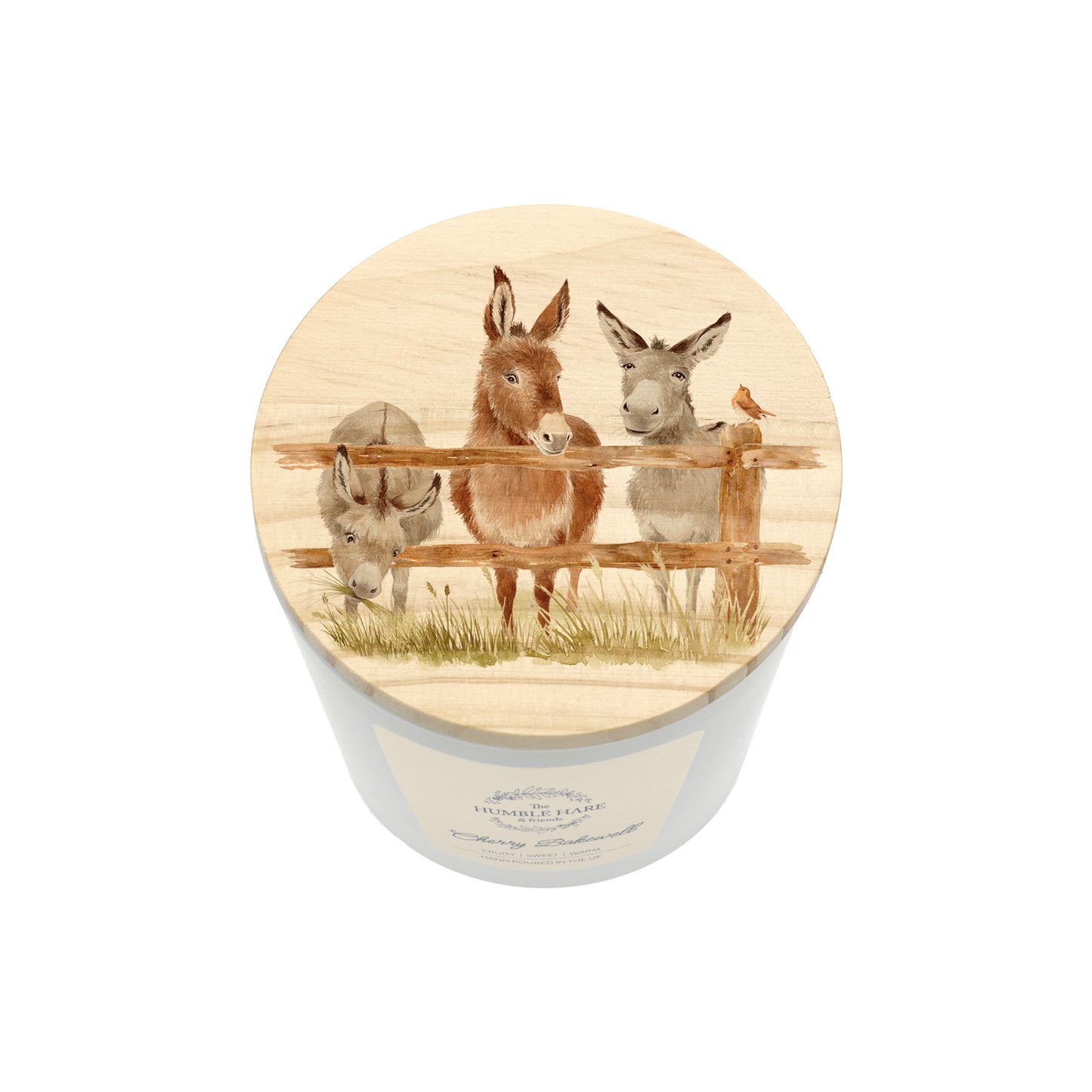 A cylindrical white candle jar with a round wooden lid with a watercolour illustration of three donkeys behind a fence and a robin, shown on a plain white background.