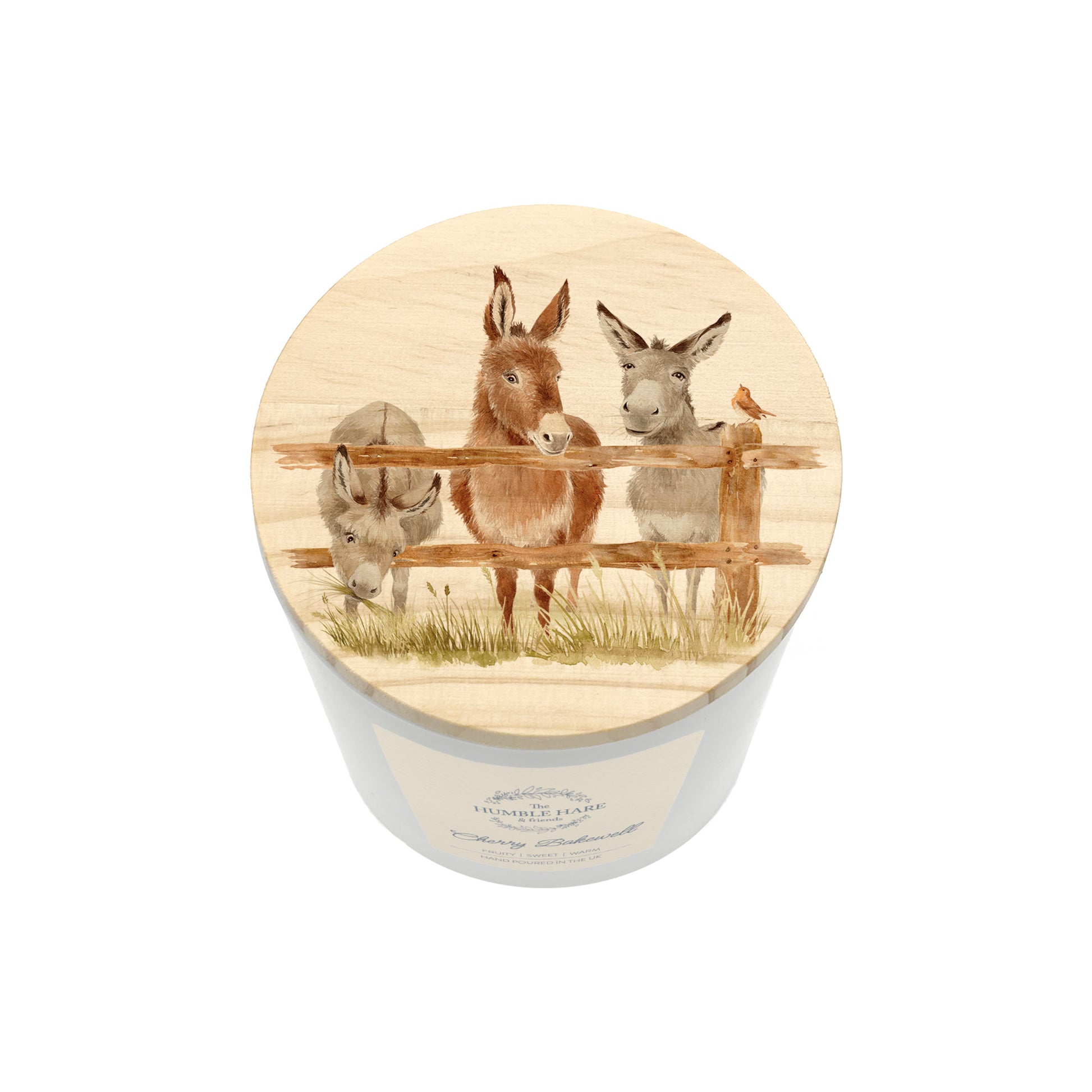 A cylindrical white candle jar with a round wooden lid with a watercolour illustration of three donkeys behind a fence and a robin, shown on a plain white background.