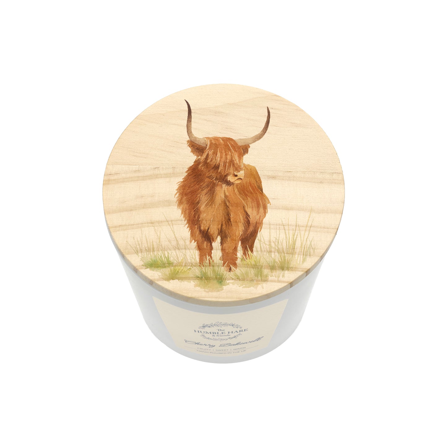 A white glass candle jar with a light wood lid with an illustration of a highland cow in a grassy field.