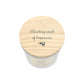 A white glass jar candle viewed from above, with a round light wood lid printed with the phrase 'Planting seeds of happiness' and a small hare silhouette.