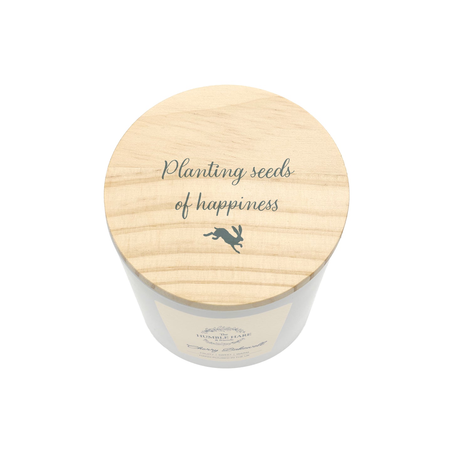 A white glass jar candle viewed from above, with a round light wood lid printed with the phrase 'Planting seeds of happiness' and a small hare silhouette.
