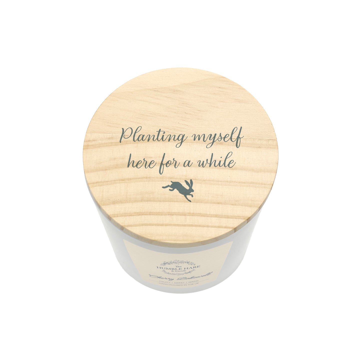 A white glass candle jar with a light wood lid with the phrase 'Planting myself here for a while' and a small leaping hare silhouette.
