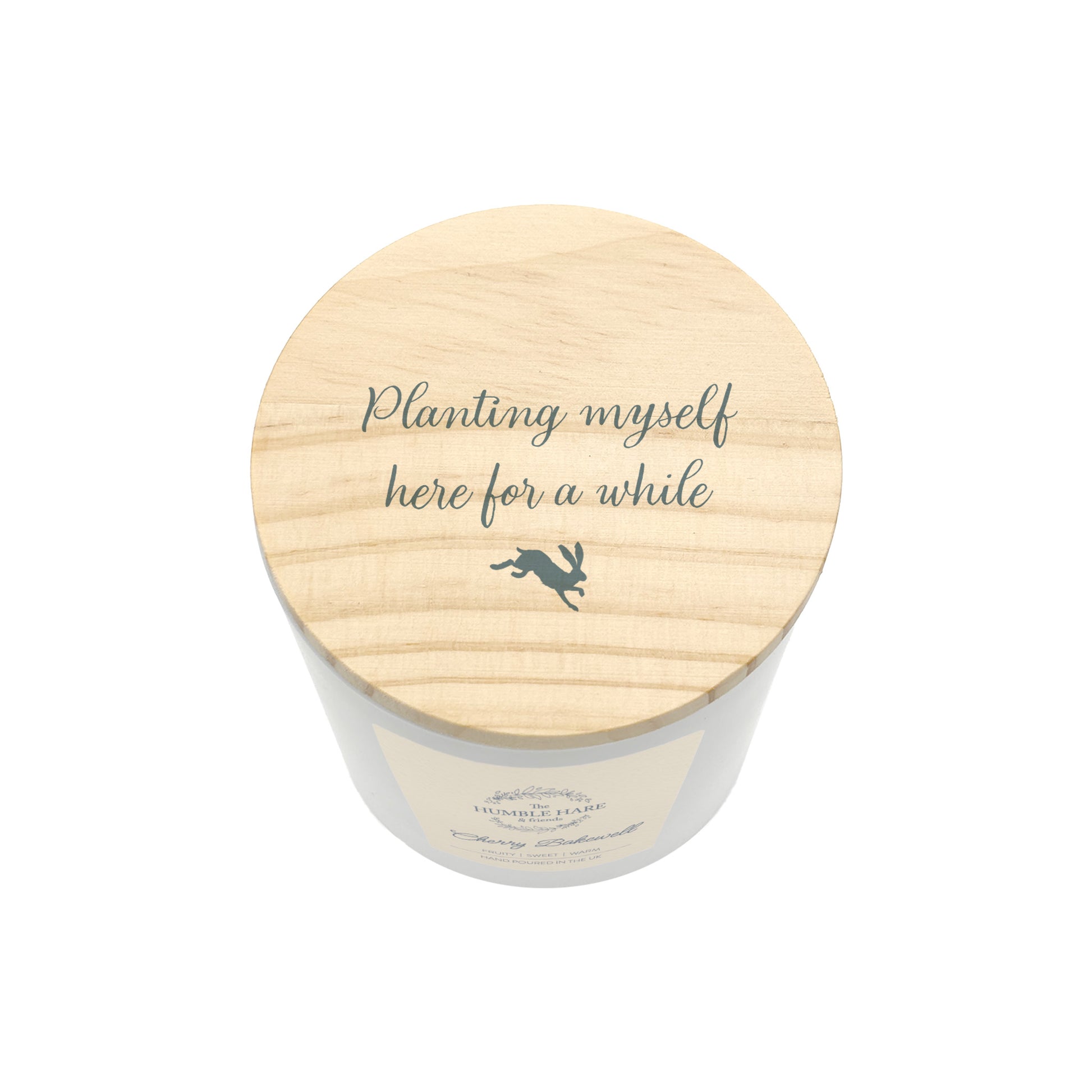 A white glass candle jar with a light wood lid with the phrase 'Planting myself here for a while' and a small leaping hare silhouette.