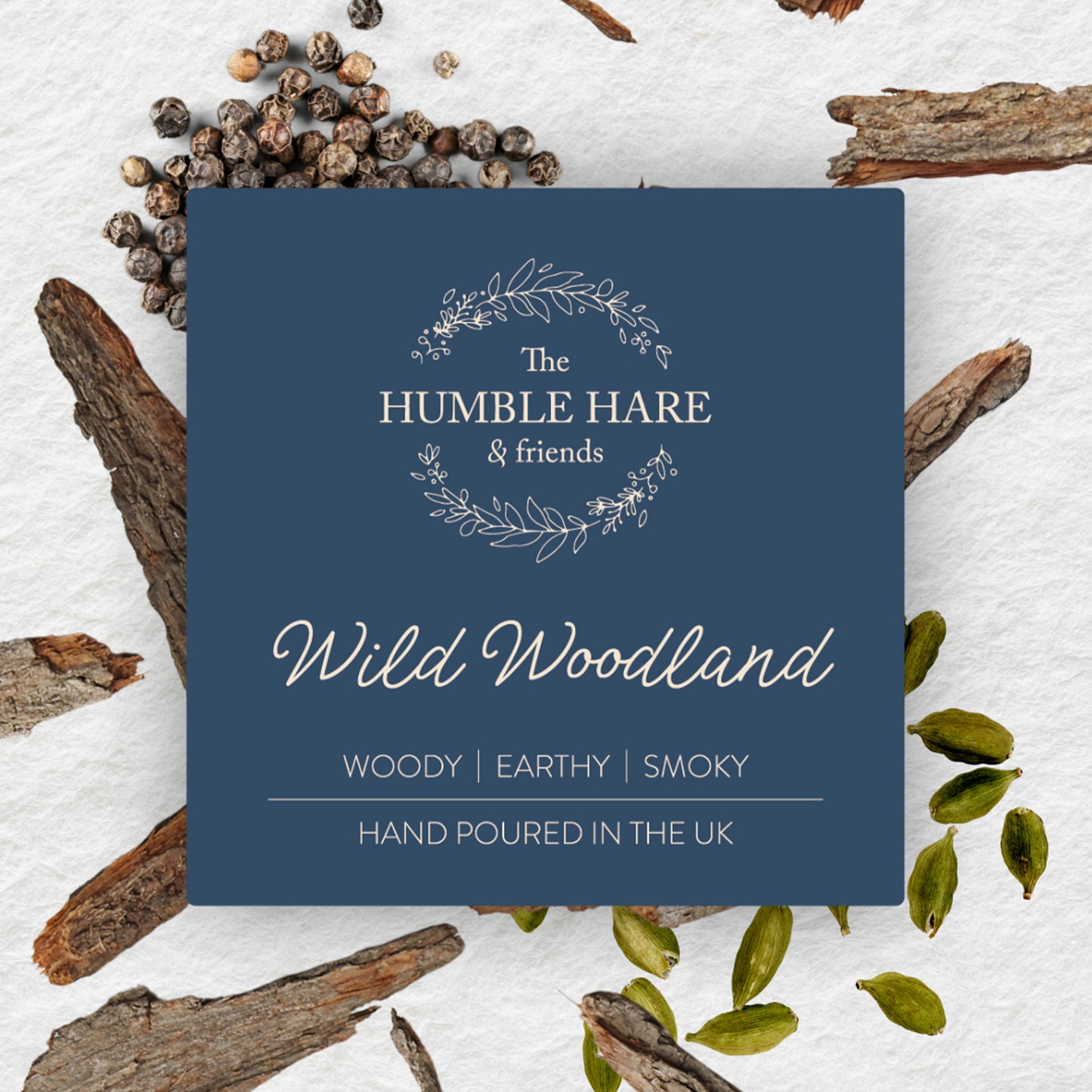a dark blue label for The Humble Hare's Wild Woodland oud and patchouli scented candle, styled on a textured paper background with bark, cardamom and pepper.