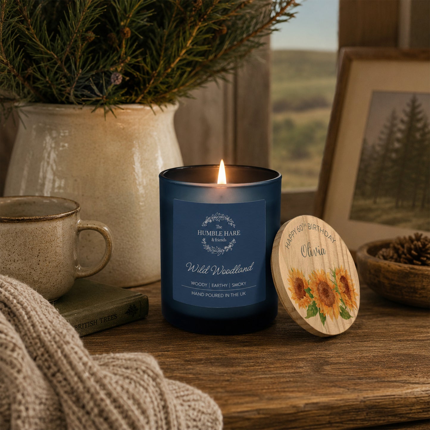A blue glass candle jar labelled 'Wild Woodland' with a lit flame, placed on a wooden table beside a personalised round wooden lid with sunflowers and a birthday message. The scene includes a mug, knit fabric, a potted evergreen, and a framed landscape photo in a cosy indoor setting.