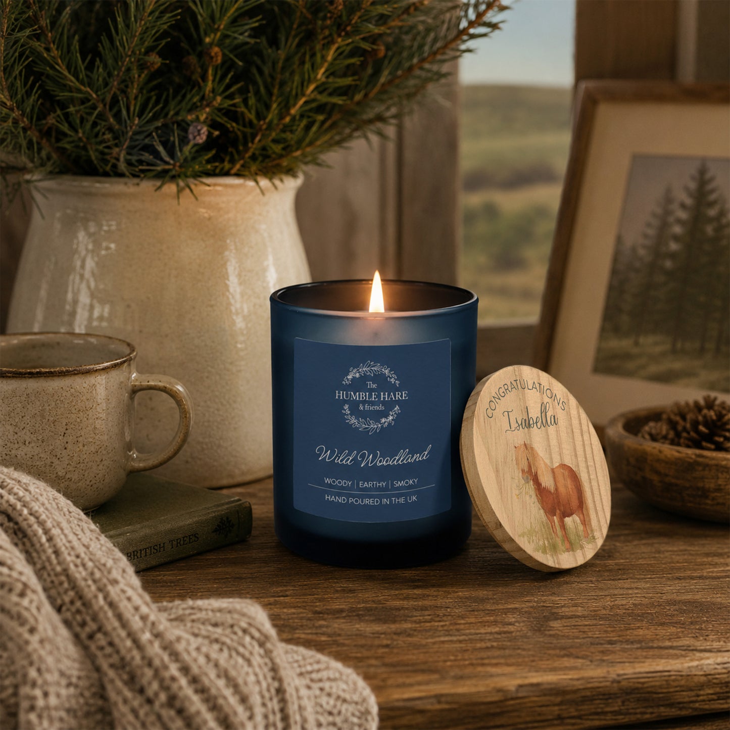 A blue glass candle jar labelled 'Wild Woodland' with a lit flame, placed on a wooden table beside a personalised round wooden lid with a Shetland pony congratulations design. The scene includes a mug, knit fabric, a potted evergreen, and a framed landscape photo in a cosy indoor setting.