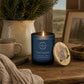 A blue glass candle jar labelled 'Wild Woodland' with a lit flame, placed on a wooden table beside a round wooden lid with a blue hydrangea. The scene includes a mug, knit fabric, a potted evergreen, and a framed landscape photo in a cosy indoor setting.