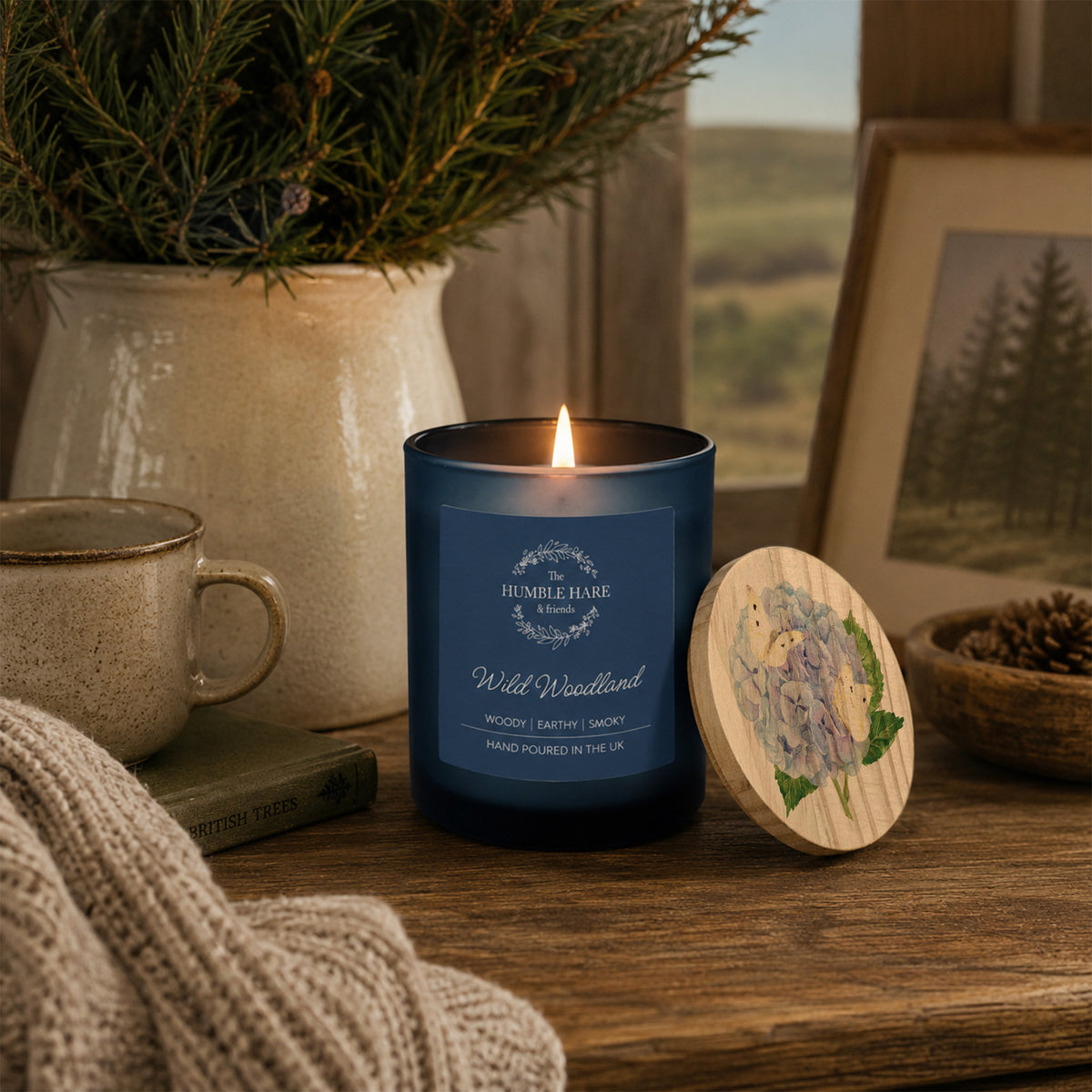 A blue glass candle jar labelled 'Wild Woodland' with a lit flame, placed on a wooden table beside a round wooden lid with a blue hydrangea. The scene includes a mug, knit fabric, a potted evergreen, and a framed landscape photo in a cosy indoor setting.