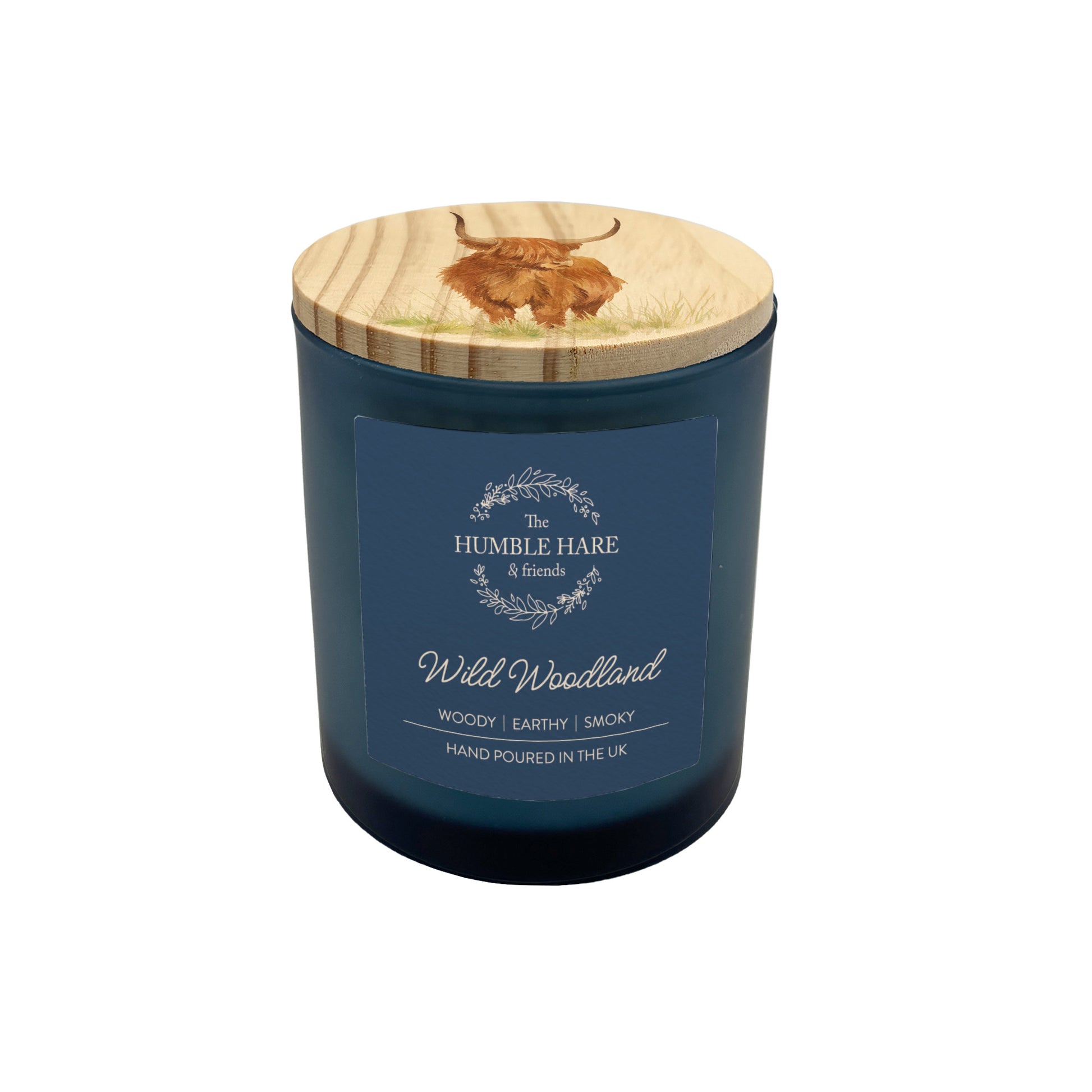 A dark blue glass jar candle with a light wood lid featuring a Highland cow, labelled 'Wild Woodland' by The Humble Hare. The candle scent is woody, earthy and smoky with oud and patchouli.