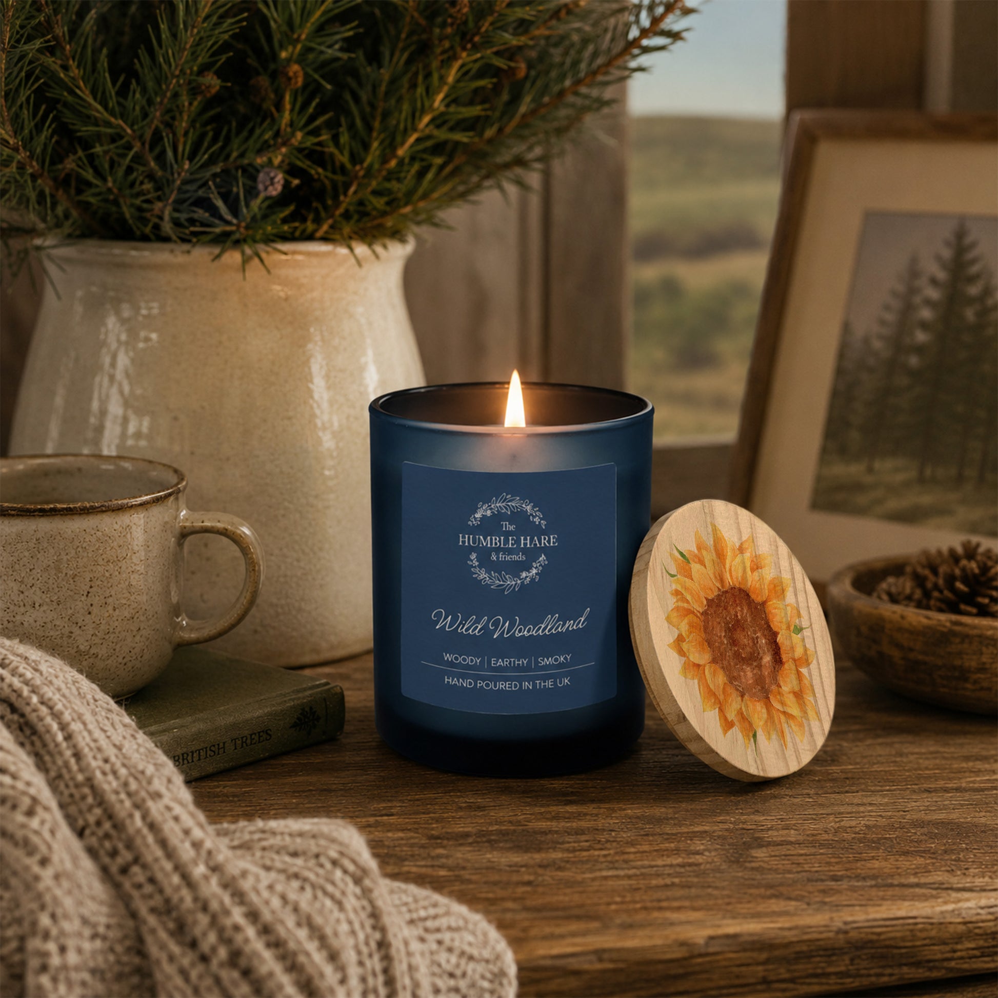A blue glass candle jar labelled 'Wild Woodland' with a lit flame, placed on a wooden table beside a round wooden lid with a watercolour sunflower design. The scene includes a mug, knit fabric, a potted evergreen, and a framed landscape photo in a cosy indoor setting.