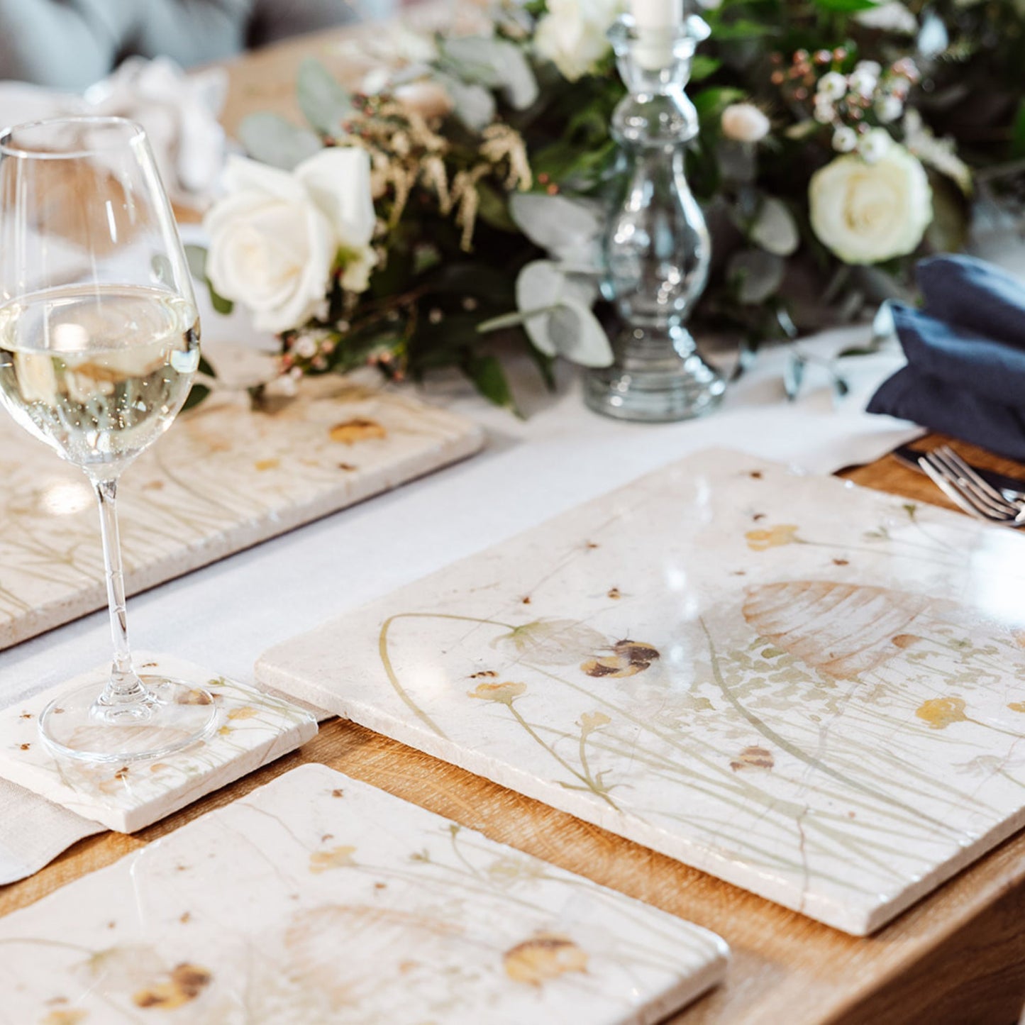 A wooden dining table set with cream marble placemats and coasters. The placemats are square and feature bees and a beehive in a buttercup meadow in a watercolour style.