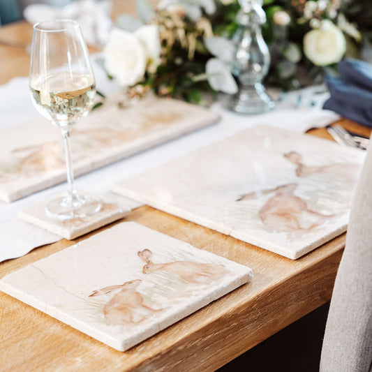A wooden dining table set with cream marble placemats and coasters. The placemats are square and feature two hares facing each other about to touch noses in a watercolour style.