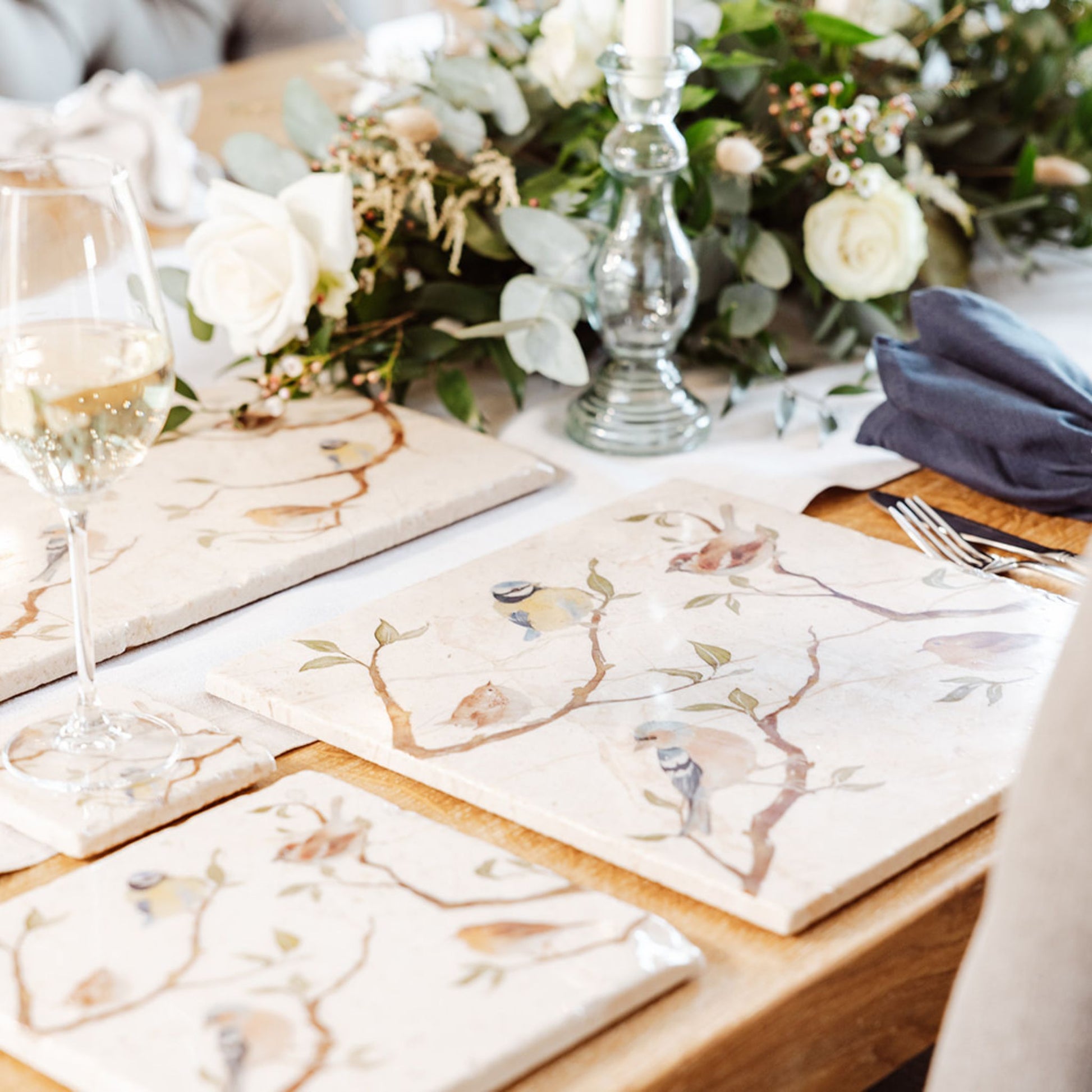 A wooden dining table set with cream marble placemats and coasters. The placemats are square and feature British garden birds on branches in a watercolour style.