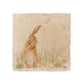 A square cream marble placemat featuring a watercolour countryside animal design of a hare in grass cleaning his ear.